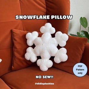 May include: A white snowflake pillow with a textured surface rests on a rust-colored pillow. The pillow is in the shape of a snowflake and is placed on a rust-colored couch. Text on the image reads "SNOWFLAKE PILLOW" and "NO SEW!!".