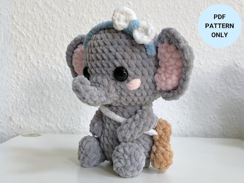May include: A gray crocheted elephant amigurumi toy with blue and white yarn accents. The elephant has a pink crocheted ear and is holding a brown crocheted object. PDF PATTERN ONLY.