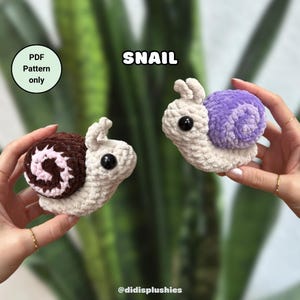 May include: Two crocheted snail plushies, one with a brown and pink shell and the other with a purple shell. The plushies are made with soft yarn and have large, black eyes. The text "SNAIL" is written above the plushies. The text "PDF Pattern only" is written in a circle in the top left corner of the image.