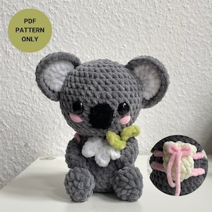 May include: A grey crocheted koala bear with a black nose, pink cheeks, and large black eyes. The koala holds a green leaf and a white flower. The image includes the text "PDF PATTERN ONLY" in a green circle.