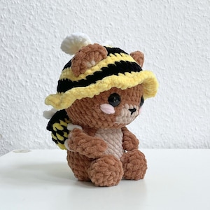 May include: A brown crocheted teddy bear wearing a yellow and black striped crocheted hat. The hat has a wide brim and is shaped like a bee.