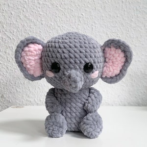 May include: A gray crocheted elephant plush toy with pink ears and black eyes. The elephant is sitting on a white surface.