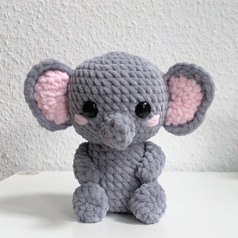 May include: A gray crocheted elephant plush toy with pink ears and black eyes. The elephant is sitting on a white surface.