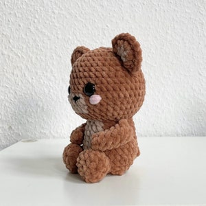 May include: A brown crocheted teddy bear with black eyes and pink cheeks. The bear is sitting with its legs crossed and its arms folded.