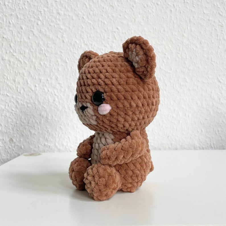 May include: A brown crocheted teddy bear with black eyes and pink cheeks. The bear is sitting with its legs crossed and its arms folded.