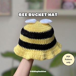 May include: A yellow and black crochet bucket hat with a bee design. The hat features black stripes and white accents on top, resembling bee wings. The text "BEE BUCKET HAT" is displayed above the hat. The hat is a PDF pattern only.