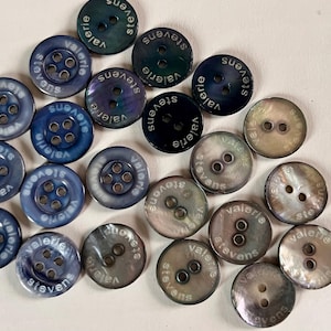 May include: A collection of round buttons in shades of blue, grey, and iridescent white. Each button features the text "valerie stevens" printed along the edge. Some buttons have four holes, while others have two holes in the center.