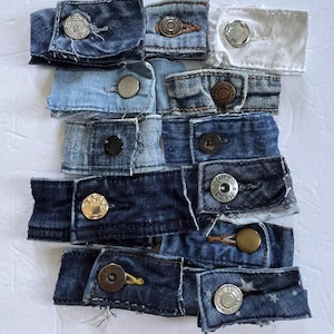 May include: A collection of denim jean button extenders in various shades of blue and white. Each extender features a button and a fabric loop, designed to add extra room to waistbands. The buttons are in silver, gold, and black.