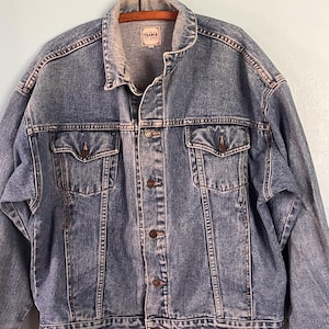 May include: A medium-wash blue denim jacket featuring a classic collar, long sleeves, and a button-front closure. The jacket includes two chest pockets with buttoned flaps and a label at the inner neckline that reads The Trader.