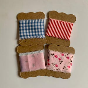 May include: Four spools of decorative ribbon in various patterns. One spool features blue and white gingham, another red and white stripes, a third pink with silver stars, and the last pink with a floral design. Each ribbon is mounted on a brown cardboard spool.