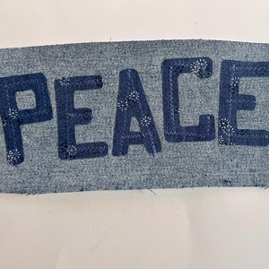 May include: A rectangular denim patch featuring the word PEACE appliqued in dark blue fabric letters. The letters are secured to the light blue denim base with a decorative white zigzag stitch pattern along the edges of each letter.