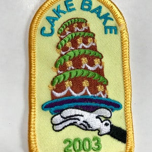 May include: An embroidered patch with a pale yellow background and a gold border. It features a multi-tiered brown cake with green icing and white accents on a purple platter held by a white gloved hand. Text reads CAKE BAKE and 2003.