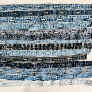 May include: An assortment of denim shirt plackets in various shades of blue, arranged horizontally. Each strip features button closures and stitching details, showcasing a range of denim washes from light to dark. The arrangement creates a textured, layered effect.
