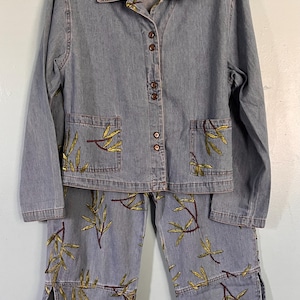 May include: A light blue denim two-piece set consisting of a long-sleeve button-down jacket and matching pants. Both pieces feature embroidered gold and dark red bamboo leaf patterns, two front pockets on the jacket, and side slits on the pants.