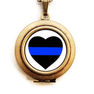 Può includere: Una collana con un ciondolo dorato a forma di cuore nero con una striscia blu al centro. Il cuore è su uno sfondo bianco.