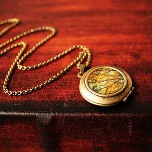 May include: A gold locket necklace with a round, hinged locket featuring a colorful image of a forest scene. The locket is on a brown leather surface.
