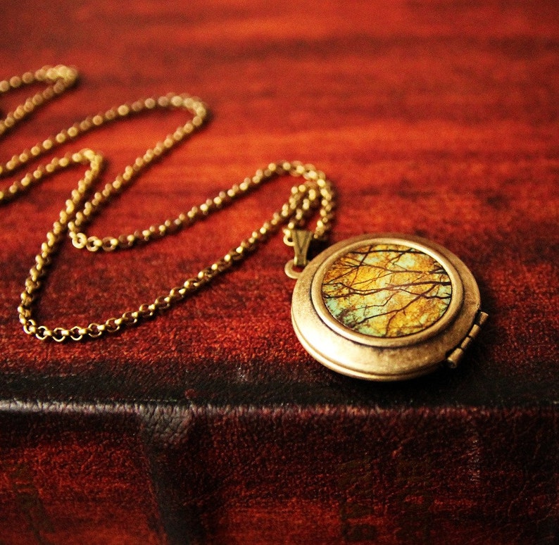 May include: A gold locket necklace with a round, hinged locket featuring a colorful image of a forest scene. The locket is on a brown leather surface.
