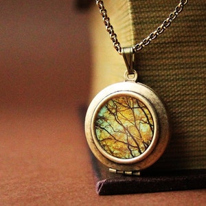 May include: A gold locket necklace with a round, brass-colored pendant featuring a nature scene of a forest with trees and branches in shades of yellow, brown, and blue.