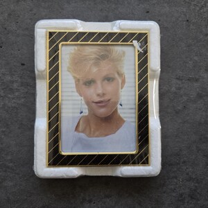 May include: A rectangular photo frame with a black and gold striped border. The frame holds a portrait of a woman with short blonde hair and earrings. The frame is set in a white protective foam.