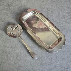 May include: A silver-toned serving spoon with a decorative floral design and a rectangular tray with a scalloped edge. The spoon has a long handle and a round bowl with cut-out patterns. The tray has a reflective surface.