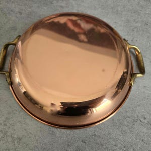 May include: A copper-colored, round pan with two brass handles. The pan has a reflective surface and a slightly raised rim. The handles are attached to the sides of the pan. The pan is likely used for cooking.