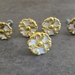 May include: Five round cabinet knobs featuring a white center with a raised gold-colored metallic floral pattern. Each knob has a circular shape with scalloped edges and a central gold dot, attached to a silver-colored metal screw post for mounting.