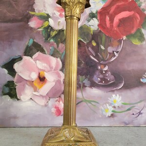 May include: A gold metal candle holder with a fluted column and a decorative base. The holder is in front of a painting of flowers.