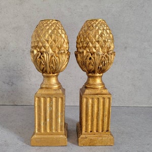 May include: Two gold-colored candle holders with a pineapple design. The holders have a fluted base and a pineapple-shaped top.