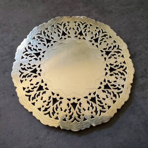 May include: A gold-colored, circular doily with an intricate, openwork design around the edge. The center is a solid, smooth surface. The doily is likely made of metal and is suitable for decorative purposes.