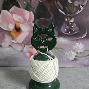 May include: A dark green cat-shaped yarn holder with a white ball of yarn. The yarn holder has a round base and a cat face design at the top. The yarn is wrapped around the holder, creating a decorative and functional item for crafters.