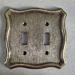 May include: A silver-toned, decorative light switch plate with a wavy, ornate border. The plate has two toggle switch openings and two screw holes.