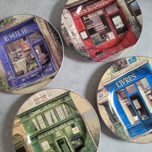 May include: Four decorative plates with images of French storefronts. Each plate features a different shop, including a bookstore with the word "LIVRES" and a restaurant named "Chez Jerome". The plates have a glossy finish and a gold rim.