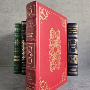 May include: A collection of antique books, with a prominent red book titled "The Devil's Dictionary" by Ambrose Bierce. The book has gold decorative accents and is part of The Franklin Library. Other books are visible in the background.