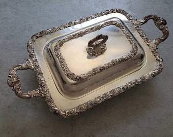 May include: Silver-plated serving dish with a rectangular shape and ornate floral and grape vine detailing. The dish has a matching lid with a decorative handle and two curved handles on the sides. A classic piece for serving food.