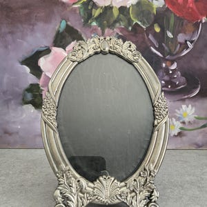 May include: An oval, silver-colored picture frame with ornate floral and scroll detailing. The frame has a dark interior and a stand for tabletop display. The design is vintage and elegant, suitable for displaying a photograph or artwork.