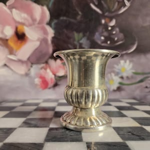 May include: A small, metallic silver-toned vase with a flared rim, a smooth cylindrical neck, and a bulbous, vertically ribbed midsection. The base is circular and features a decorative, scalloped edge, providing a stable foundation for the vessel.