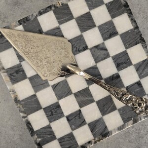 May include: A silver cake server with an ornate floral design rests on a black and white checkered marble surface. The server's handle features detailed carvings, and the blade is triangular. The marble base has a gray border.