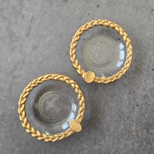 May include: Two small, circular glass dishes featuring a clear, transparent center and a textured, metallic gold rim designed with a braided rope pattern. Each dish includes a single, small, solid gold-colored tab extending from the outer edge.