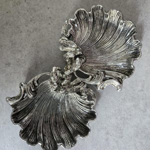 May include: A silver-toned, ornate decorative dish with a seashell design. The dish features a detailed, textured surface with a swirling, fan-like pattern. The dish is made of metal and has a vintage aesthetic.