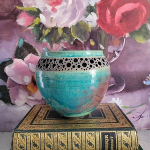 May include: A teal ceramic bowl with a decorative cut-out design around the rim. The bowl is sitting on top of two green and gold books. The books have the titles "J. Fouche Memoirs" and "Ernest Hemingway The First Forty-Nine Stories".