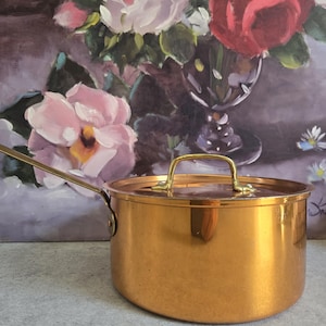 May include: A copper saucepan with a lid and a gold-colored handle. The pot is round with a shiny finish. The background features a floral still life painting with roses and other flowers.