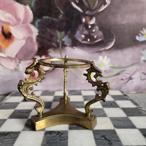 May include: A brass-colored decorative stand with a circular top and three dragon-shaped supports. The stand is designed to hold a small sphere or object. The base is a square shape.