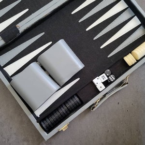 May include: A gray backgammon game set with a black felt board, white and gray playing pieces, and two dice. The set is open and ready to play.