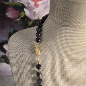 May include: A beaded necklace with dark purple and gold accents. The necklace features large, round, dark purple beads strung together with a gold clasp and smaller gold beads. The necklace is displayed on a mannequin.