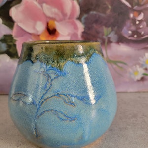 May include: A small, light blue ceramic vase with a green and brown glaze. The vase has a delicate, hand-painted design of a leafy branch.