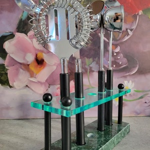 May include: A set of five silver cocktail tools with black handles, including a cocktail shaker, a strainer, a muddler, and two spoons, displayed in a green marble stand with a clear acrylic top.