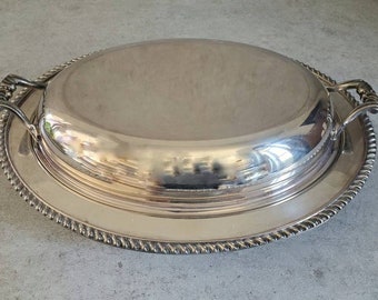 May include: A silver-colored, oval serving dish with a lid and handles. The dish sits on a matching oval tray with a rope-like border. The lid has a curved shape, and the handles are ornate.