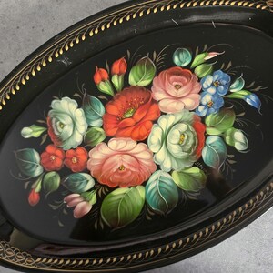 May include: Oval black tray with a floral design. The tray features a gold trim and a hand-painted bouquet of red, pink, white, and blue flowers with green leaves. The tray is a decorative item.