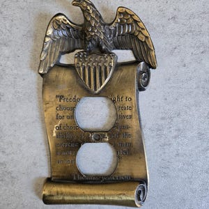 May include: A decorative brass-toned electrical outlet cover featuring an eagle perched above a shield with stars and stripes. The cover includes a scroll with text from Thomas Jefferson. The eagle has detailed feathering.