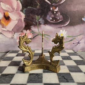 May include: A small, ornate brass display stand with a central vertical support and decorative, scroll-like side elements. The stand is positioned on a checkered black and white surface, with a floral painting in the background. The stand is approximately 4 inches tall.